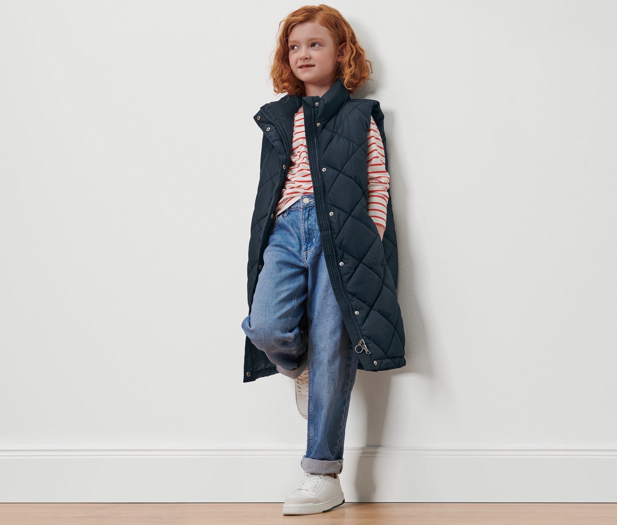 Kızıl saçlı bir kız duvara yaslanmış, uzun, kapitone çocuk yeleği giyiyor.