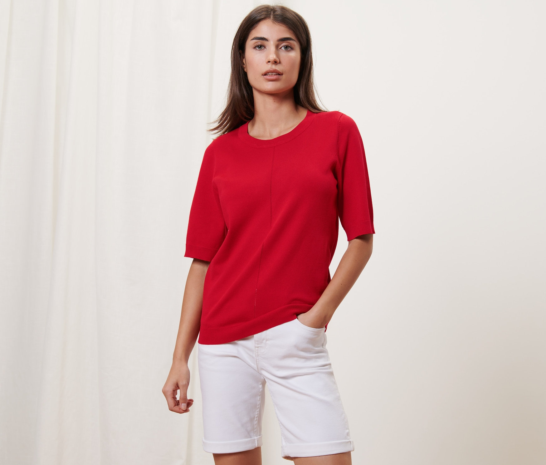 Kadın kırmızı ince örgülü kazak ve beyaz şortla poz veriyor.