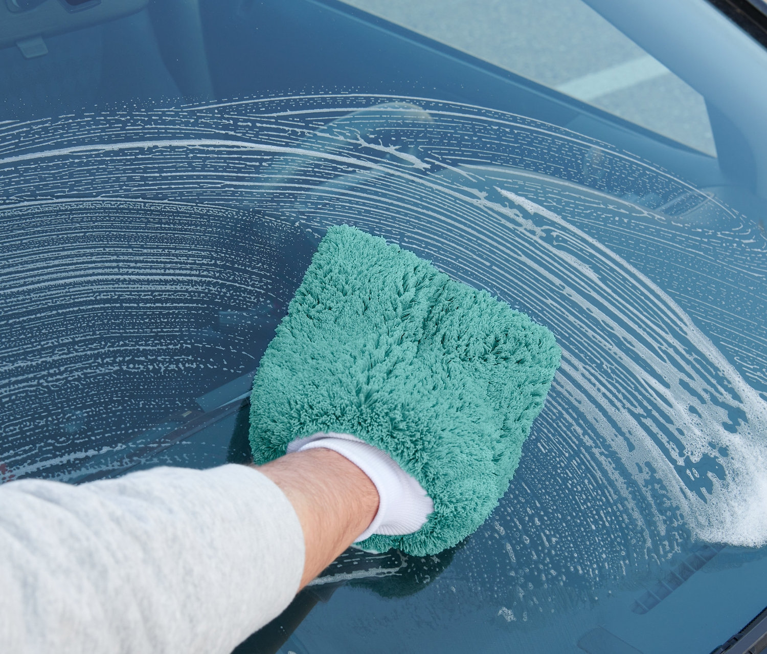 Adam bir araba yıkama eldiveni ile arabanın ön camını siliyor.