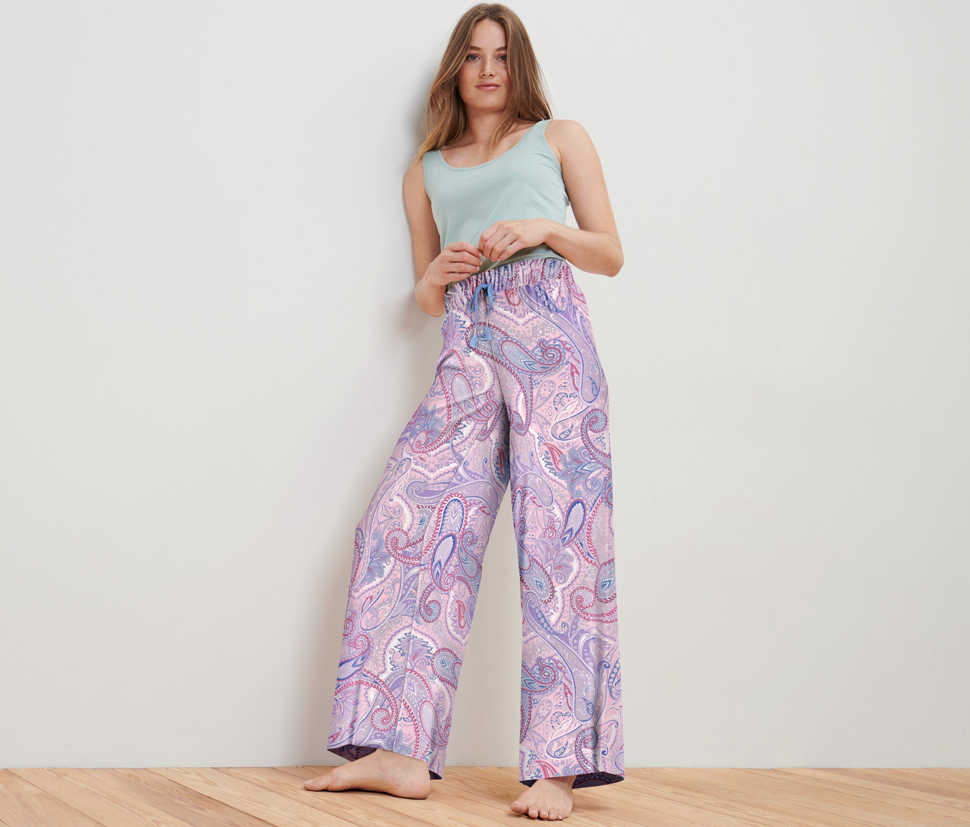 Kadın stüdyoda poz veriyor, üzerinde paisley desenli dokuma rahat pantolon ve açık yeşil bir bluz var.