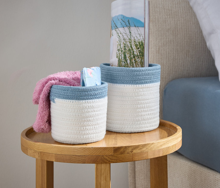 Mavi kenarlı iki beyaz örgü sepet, yatağın yanındaki ahşap bir masanın üzerinde duruyor.