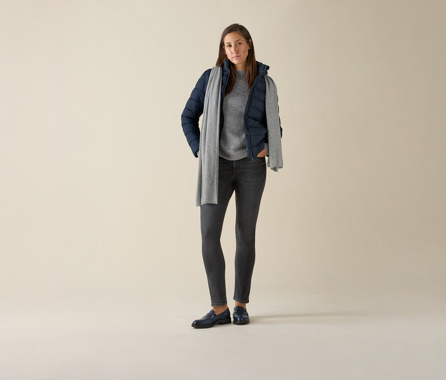 Ayakta duran bir kadın gri denim, gri bir kazak, lacivert bir ceket ve gri bir eşarp ile Slim-Jeans – Fit »Emma« giyiyor.