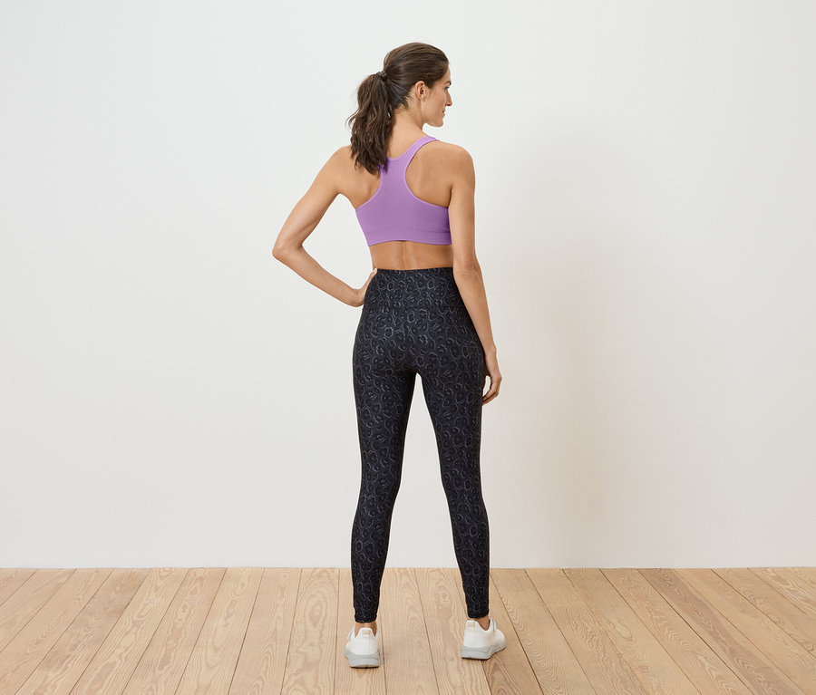 Kadın arkası dönük duruyor, mor bir sporcu sütyeni ve siyah tayt giyiyor.