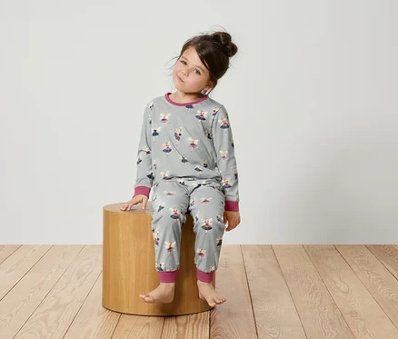 Kız çocuğu tahta bir taburede oturuyor ve üzerinde balerin desenli gri bir pijama var.