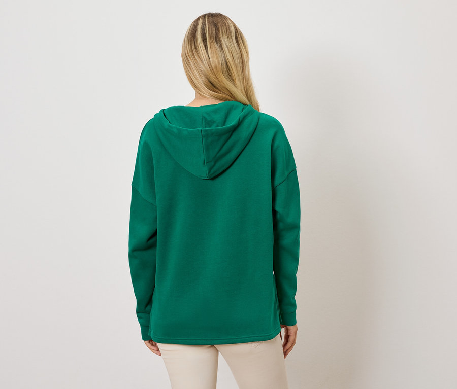 Sırtı kameraya dönük bir kadın, yeşil kapüşonlu sweatshirt ve açık renk pantolon giyiyor.