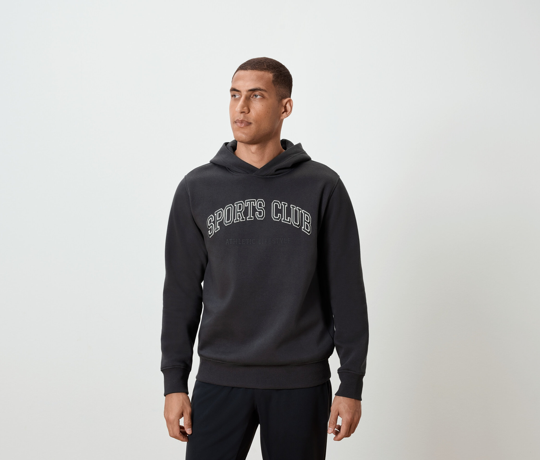 Adam koyu gri renkte, üzerinde SPORTS CLUB yazan kapüşonlu bir sweatshirt giyiyor.