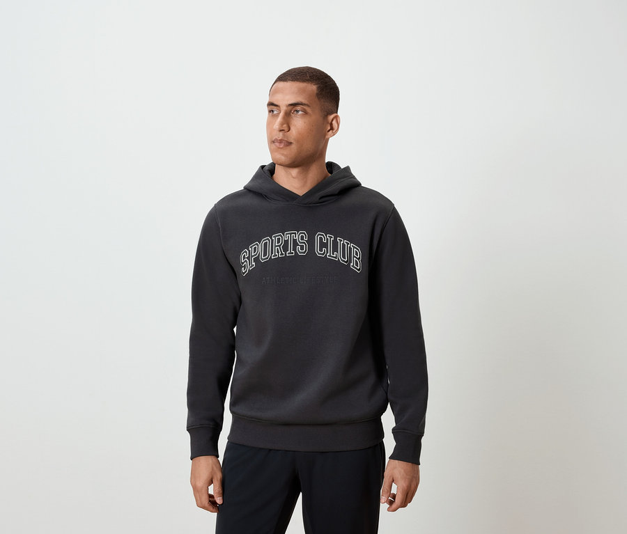 Adam koyu gri renkte, üzerinde SPORTS CLUB yazan kapüşonlu bir sweatshirt giyiyor.