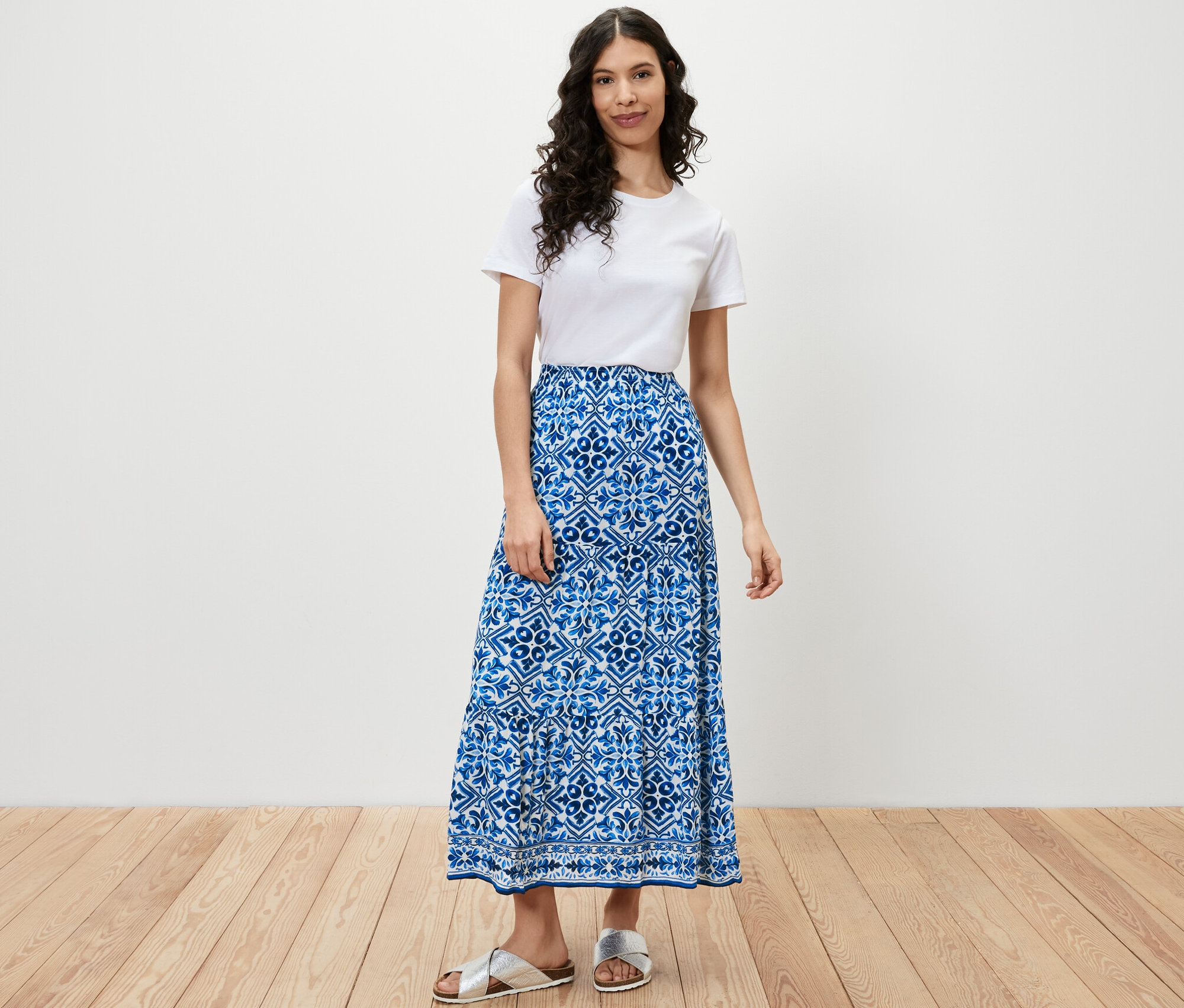 Kadın ayakta duruyor, beyaz tişört ve baskılı dokuma etek giyiyor.