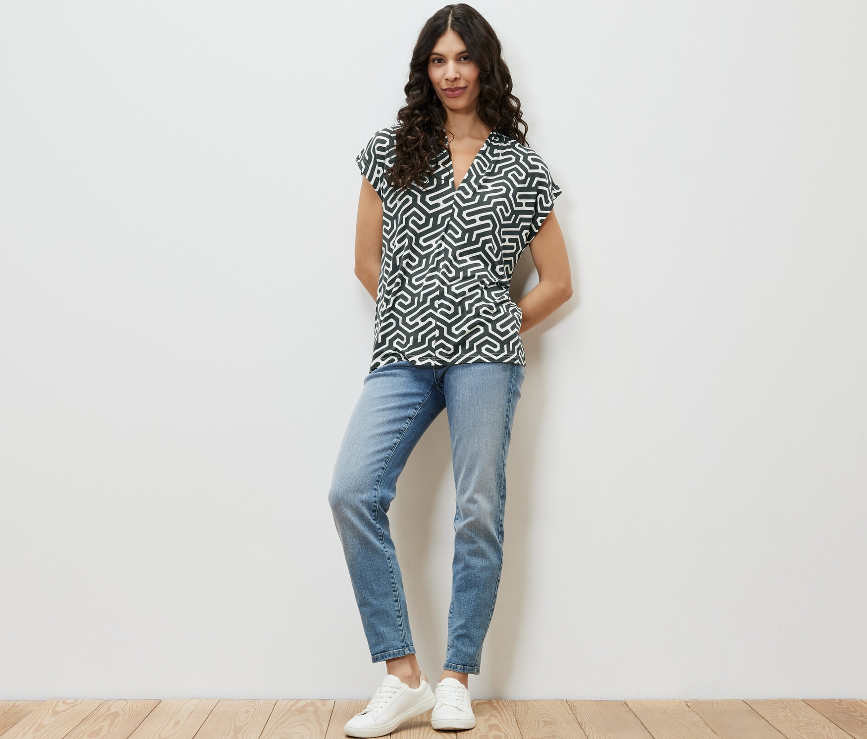 Bir kadın ayakta duruyor ve yeşil V yakalı bluz, mavi kot pantolon ve beyaz spor ayakkabı giyiyor.