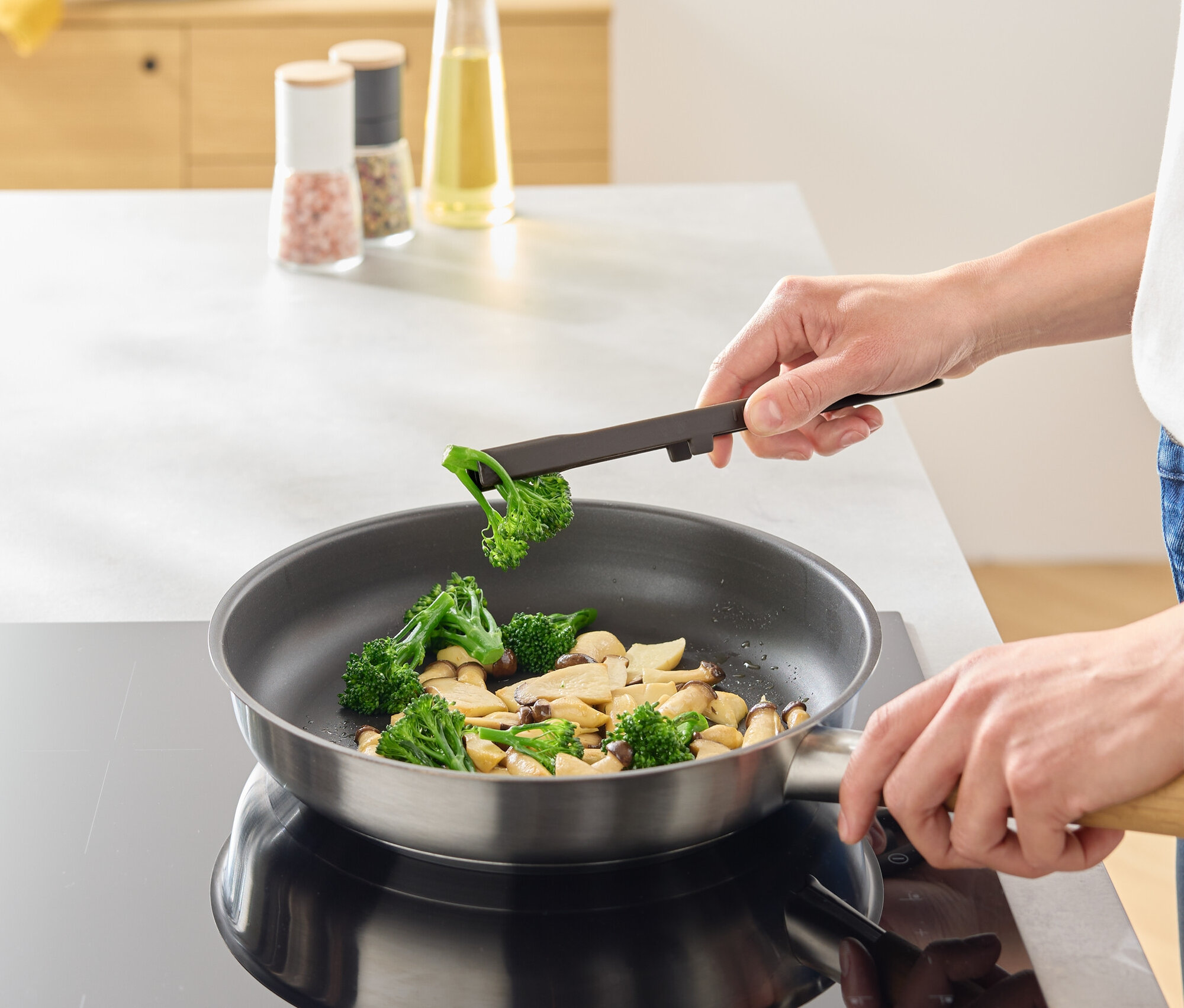 Bir kişi maşa yardımıyla bir tavaya brokoli ekliyor.
