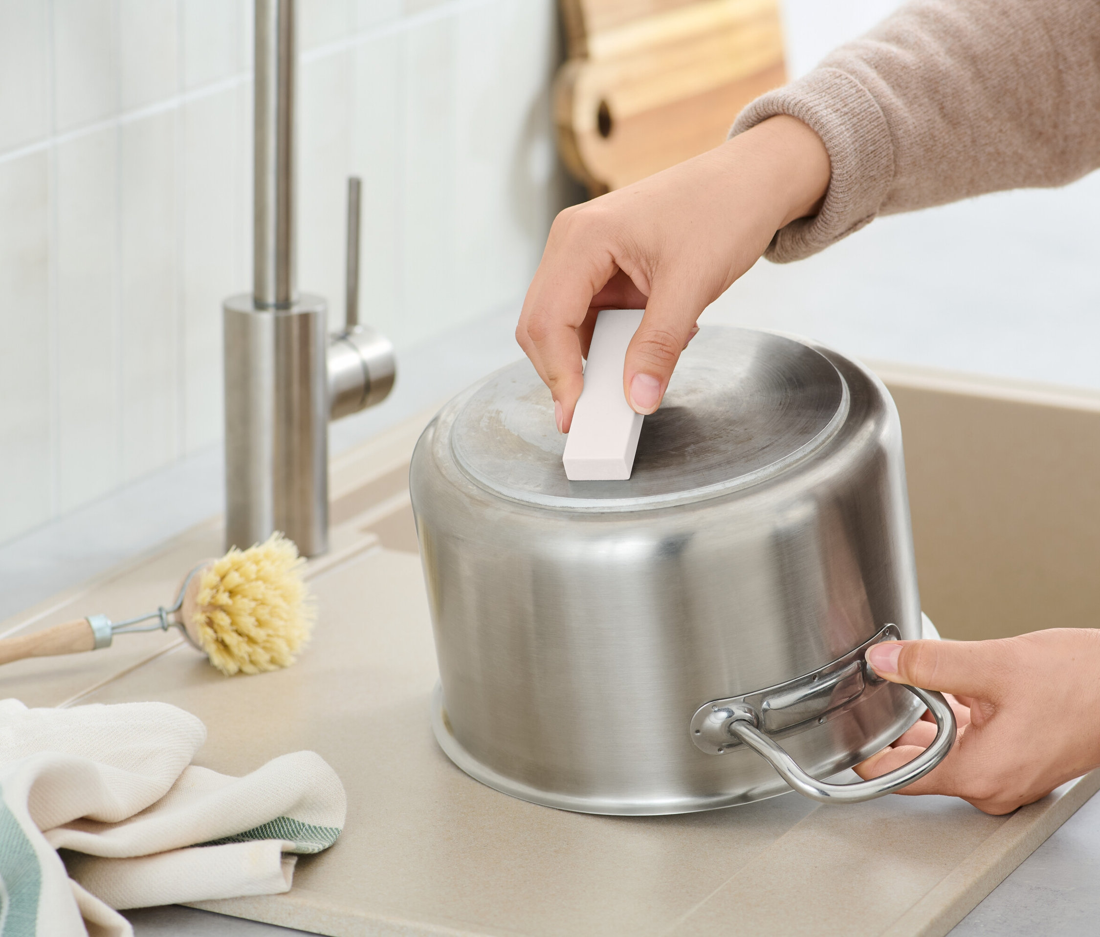 Bir kişi, lavaboda paslanmaz çelik bir tencereyi temizleme süngeriyle temizliyor, yanında bir fırça ve bir bez var.