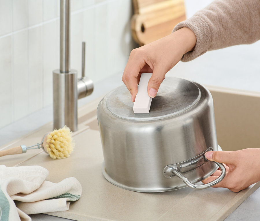 Bir kişi, lavaboda paslanmaz çelik bir tencereyi temizleme süngeriyle temizliyor, yanında bir fırça ve bir bez var.