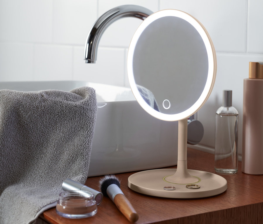 Kahverengi bir dolabın üzerinde gri bir havlu, lavabo ve kozmetik ürünlerinin yanında ışıklı bir LED Makyaj Aynası duruyor.