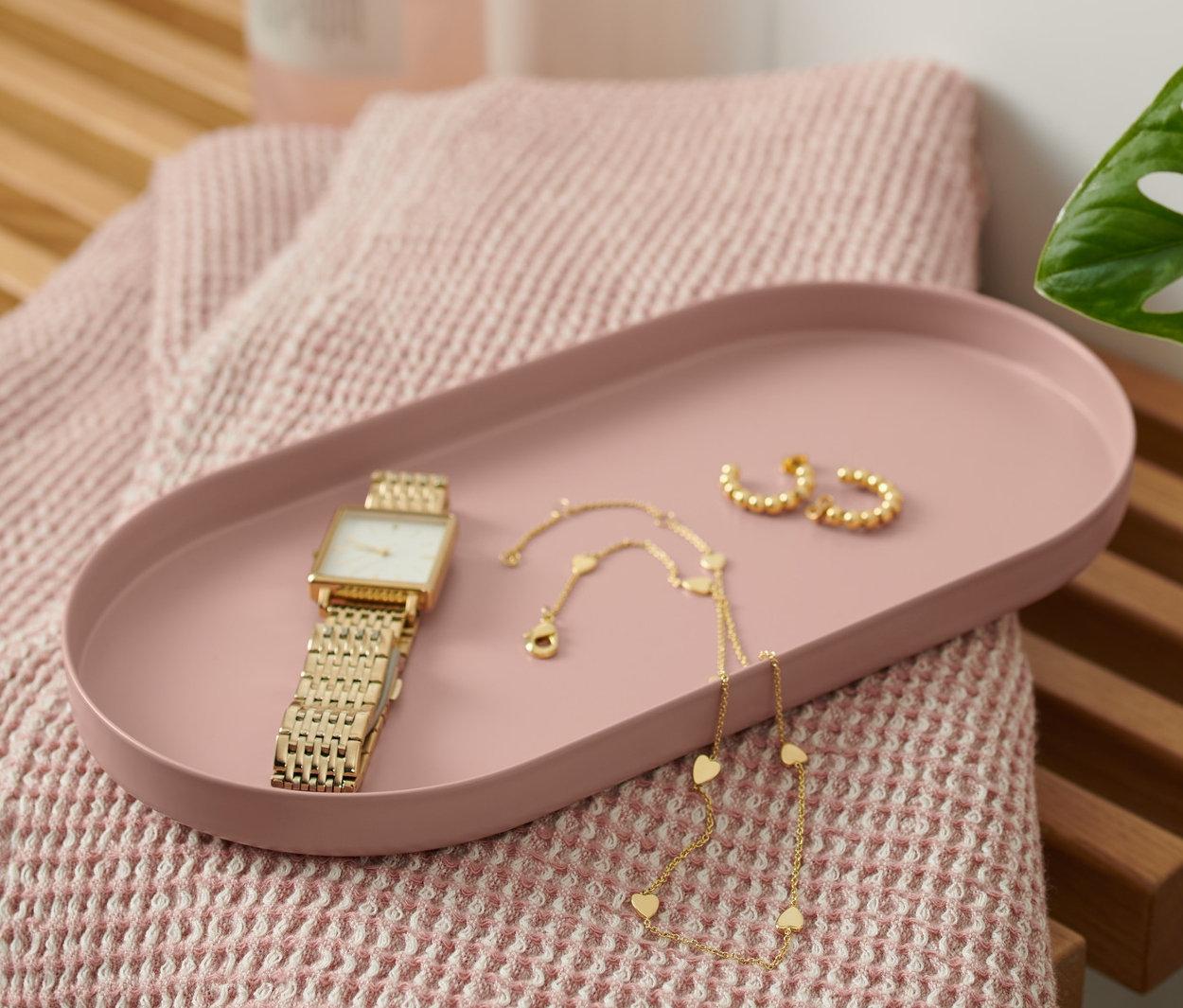 Ahşap bir bank üzerinde bakır rengi metal tepsi, saat, kolye ve küpeler bulunmaktadır. Tepsinin yanında pembe bir havlu ve pompalı bir şişe bulunmaktadır.