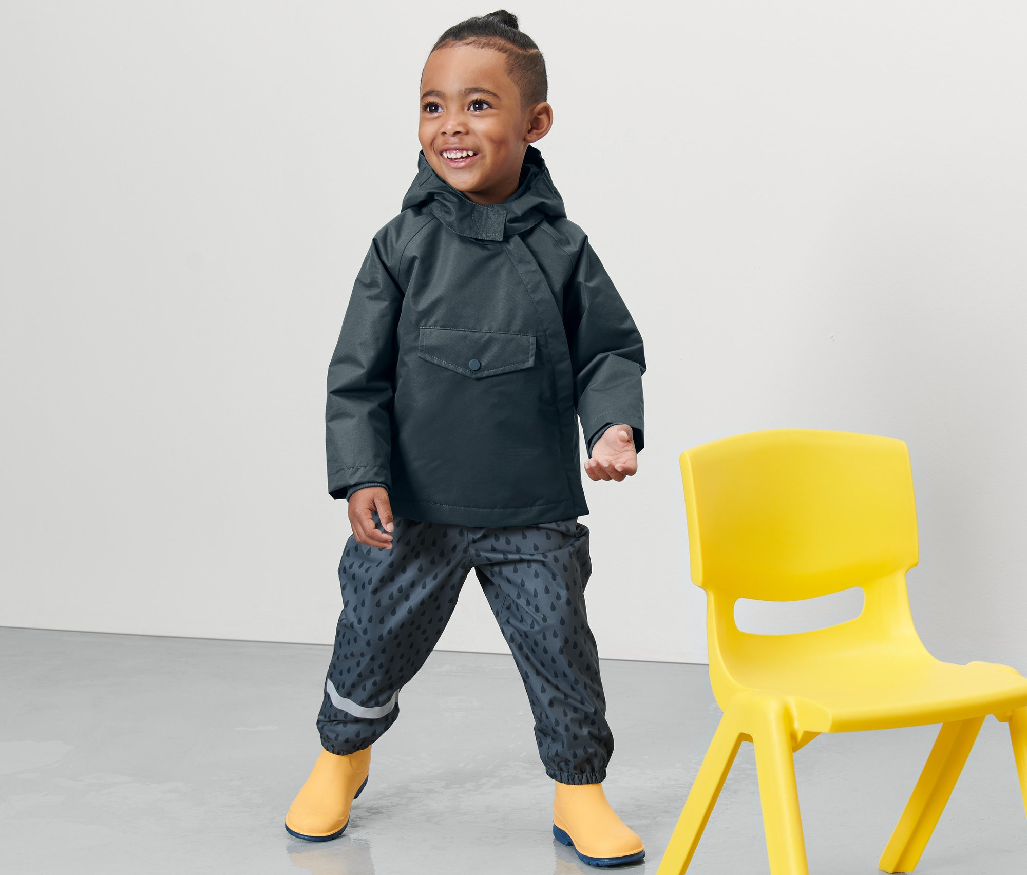 Bir çocuk sarı bir sandalyenin yanında duruyor ve Kayrak Mavisi renginde Küçük Çocuk Termal Yağmurluk giyiyor.