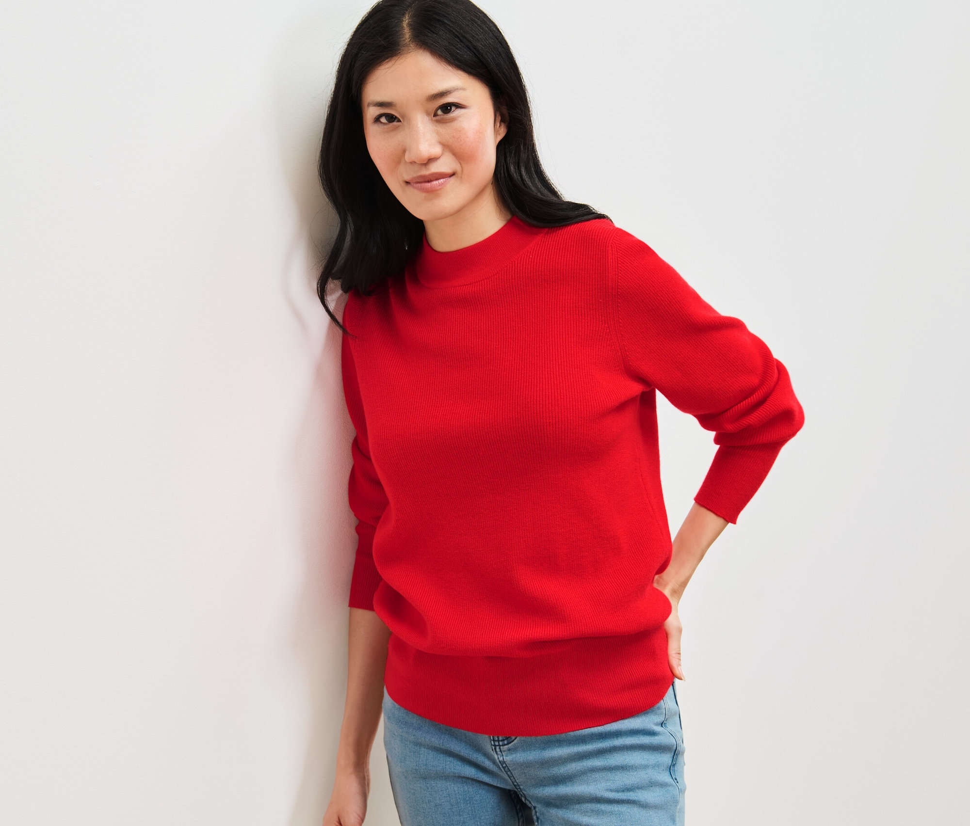 Duvara yaslanmış, kırmızı ince örgü kazak ve kot pantolon giyen bir kadın.