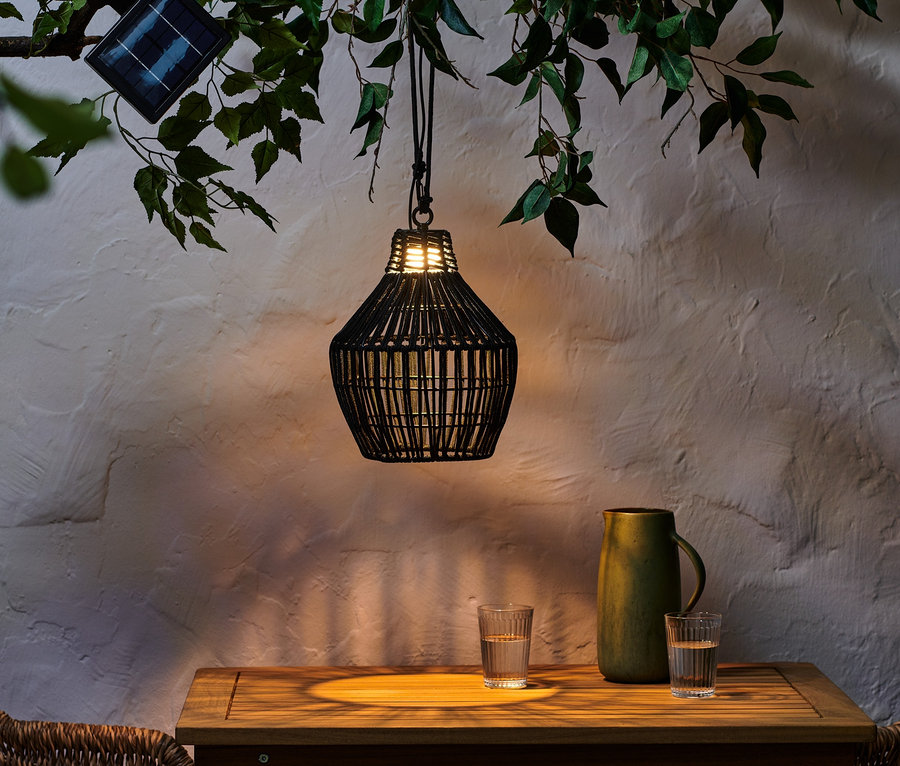 Siyah, tekstil bantlı güneş enerjili sarkıt lamba, bardakların ve yeşil bir sürahinin bulunduğu masanın üzerinde bir daldan sarkıyor.