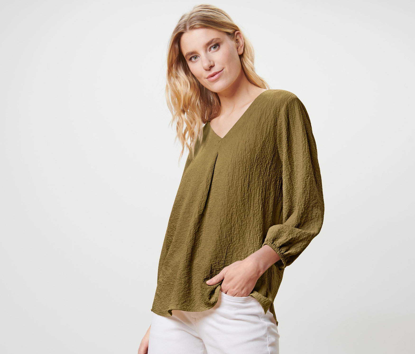 Sarışın bir kadın zeytin yeşili bir tunik bluz ve beyaz pantolonla poz veriyor.