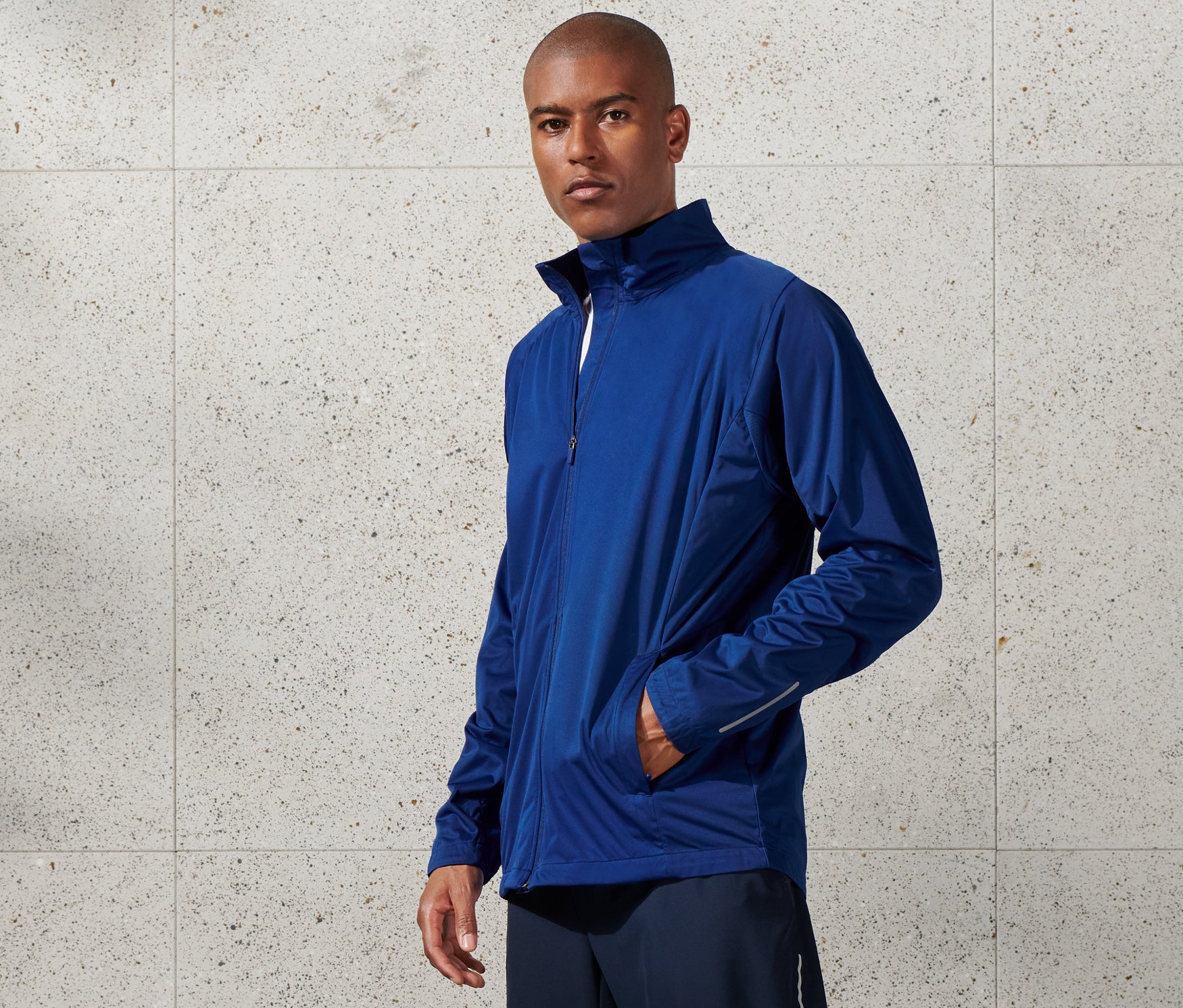 Adam mavi rüzgar korumalı fonksiyonel ceket, şort ve beyaz spor ayakkabı giyiyor.