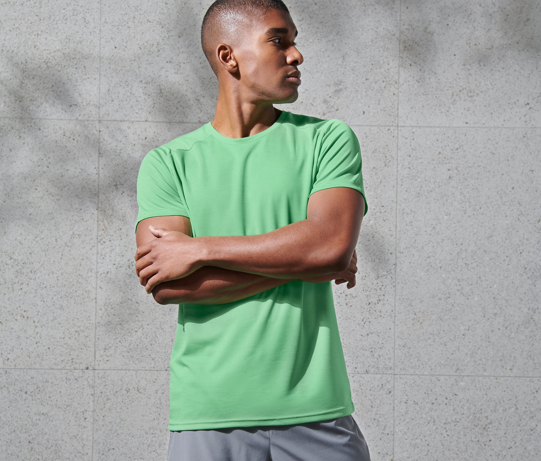 Kollarını kavuşturmuş bir adam yeşil fonksiyonel tişört ve gri şort giyiyor.