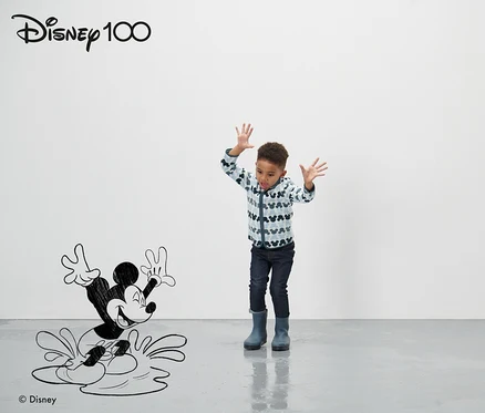 Bir çocuk çizgi film Mickey Mouse figürünün önünde duruyor ve ellerini sallıyor. Mavi Küçük Çocuk Polar Ceket »Micky« giyiyor.
