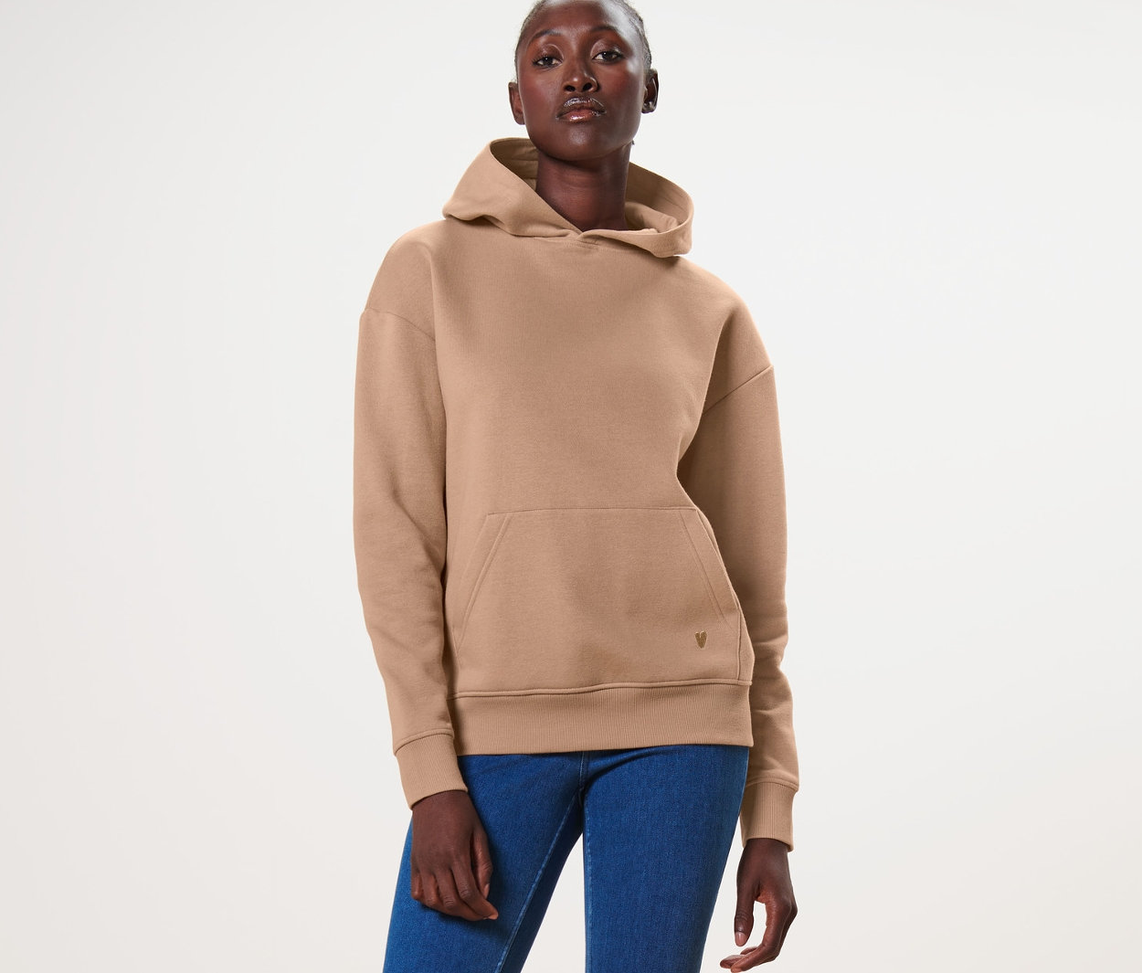 Kadın ayakta duruyor ve bej rengi bir kapüşonlu, mavi kot pantolon ve beyaz spor ayakkabı giyiyor.