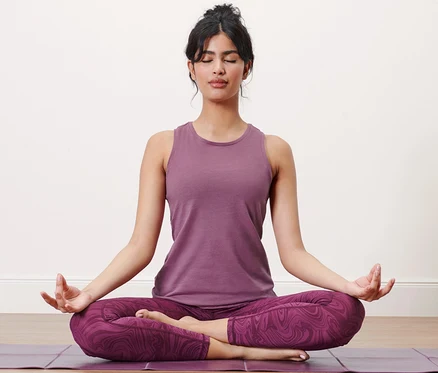 Kadın bir matın üzerinde lotus pozisyonunda oturuyor ve eflatun rengi bir spor üstü giyiyor.