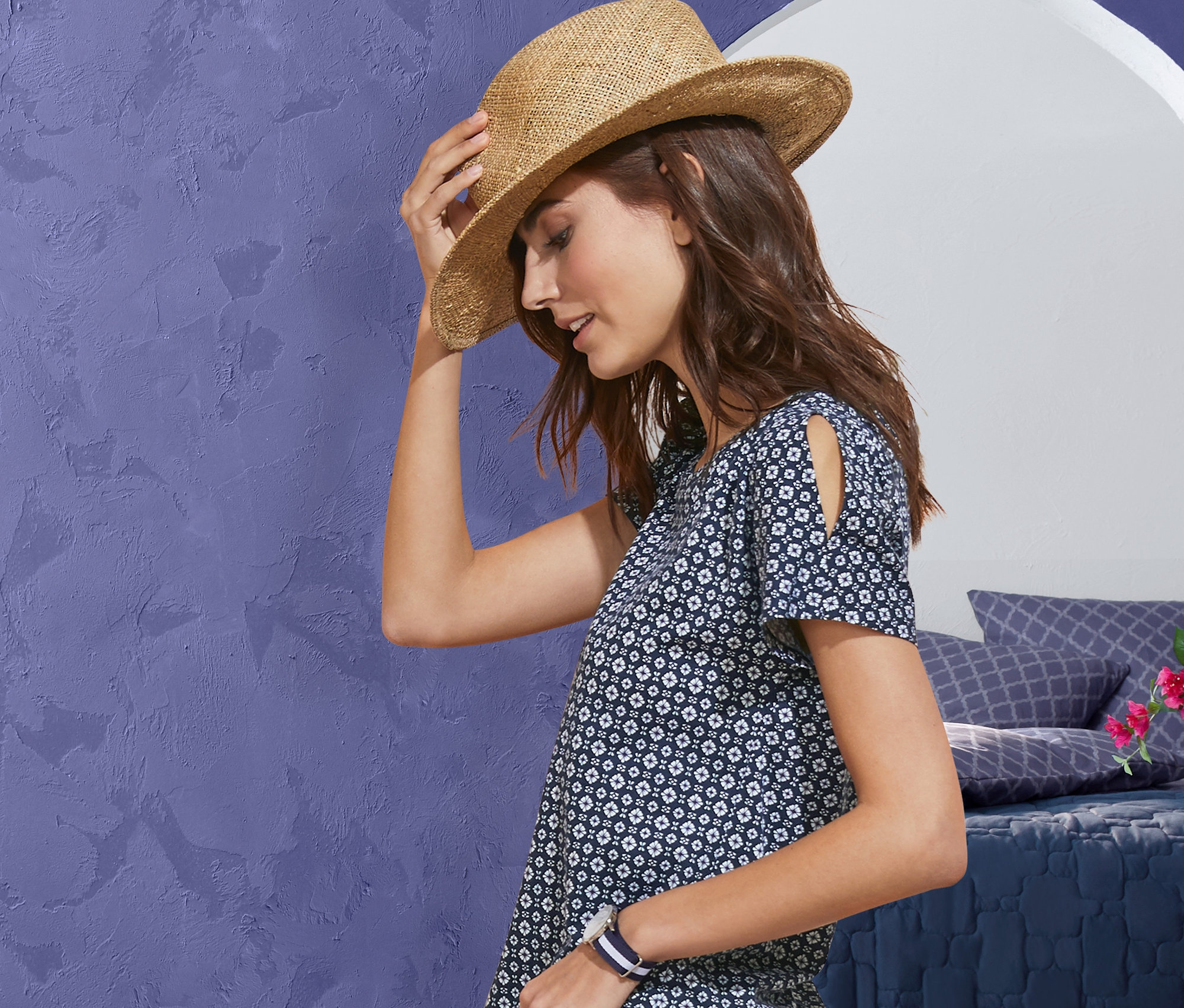 Kadın bir hasır şapka tutuyor, desenli bir tişört ve 7/8 boy stretch jean pantolon giyiyor. Arka planda yatak örtüsü, kapitone yatak örtüsü ve puf görülüyor.