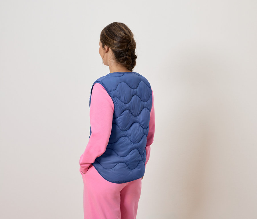 Sırtı kameraya dönük bir kadın, pembe pijamasının üzerinde mavi, kapitone bir yelek giyiyor.