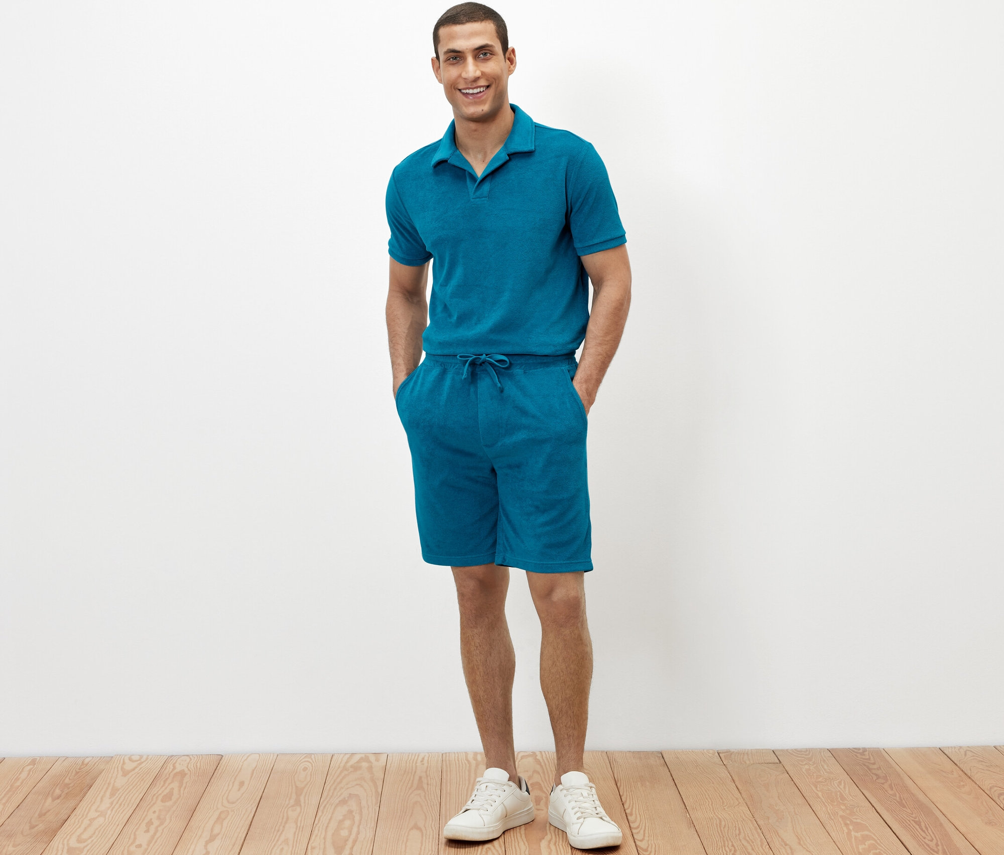 Adam, petrol rengi havlu şortunun ceplerinde elleriyle ayakta duruyor.