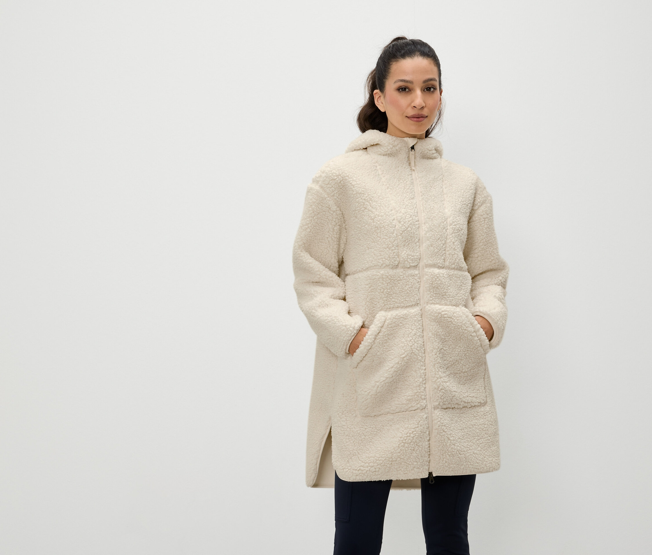Kadın, krem rengi, fermuarlı, kapüşonlu, astarlı bir montun ceplerinde elleriyle duruyor.