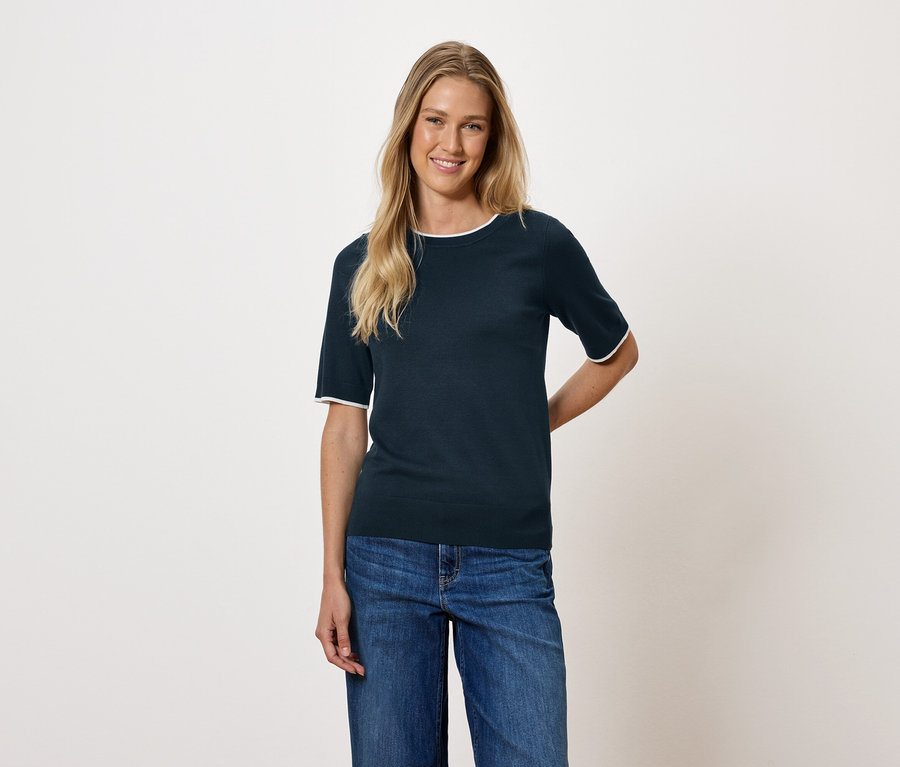 Sarışın saçlı bir kadın, lacivert kısa kollu bir bluz ve kot pantolon giyiyor.