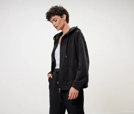 Kısa kıvırcık saçlı bir kadın, siyah kapüşonlu bir sweatshirt, siyah eşofman altı ve beyaz bir bluz giyiyor.