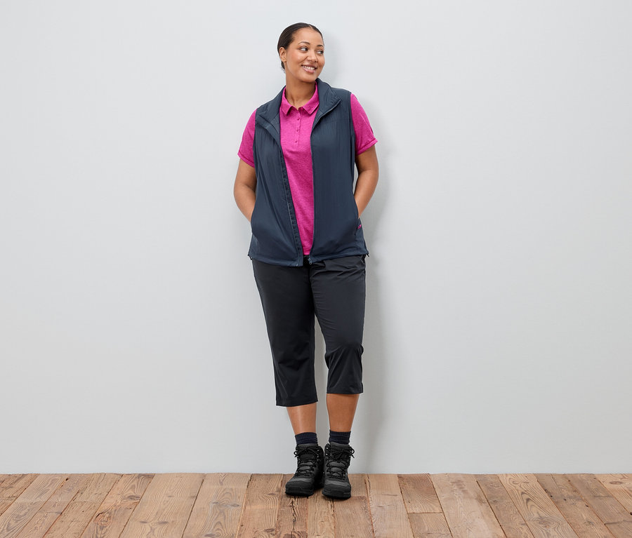 Kadın elleri cebinde bir duvarın önünde duruyor. Üzerinde pembe bir polo yakalı tişörtün üzerine lacivert bir yelek, siyah pantolon ve botlar var.