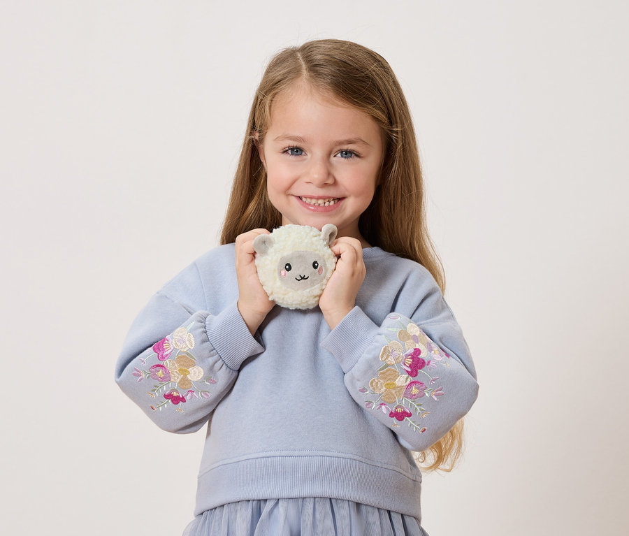 Gülümseyen küçük bir kız, önünde peluş bir koyun tutuyor. Kollarında çiçek işlemeleri olan mavi bir kazak giyiyor.