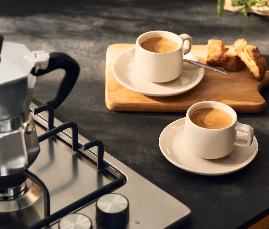 Gazlı ocakta gümüş renkli moka pot, yanında iki açık renkli kahve fincanı ve altlıkları ile ahşap bir tahta üzerinde biscotti.