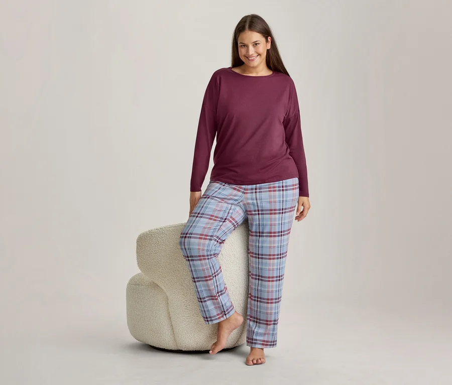 Bordo uzun kollu tişört ve açık mavi, kırmızı, beyaz ekose pijama pantolonu giyen gülümseyen kadın, bir ayağını tüylü beyaz bir tabureye koymuş duruyor.