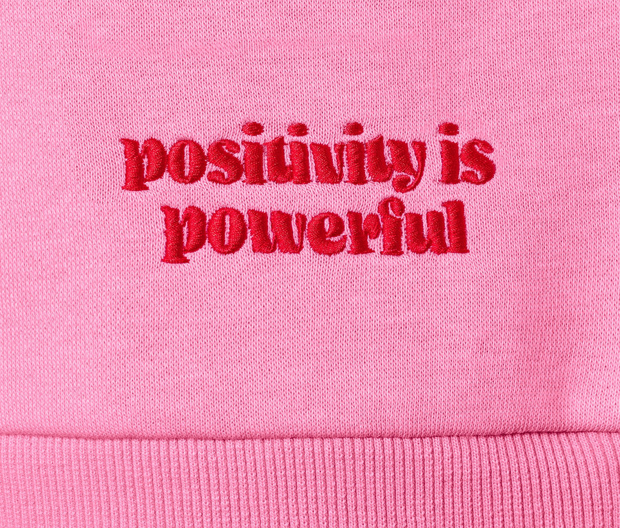 Pembe bir sweatshirt'ün üzerinde kırmızı renkte "positivity is powerful" yazısının detaylı görüntüsü.