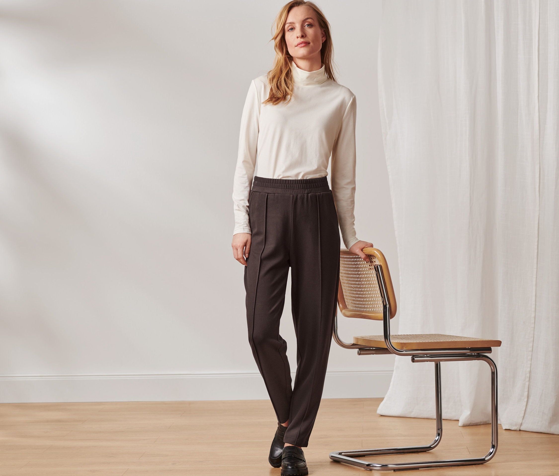 Kadın bir sandalyenin yanında poz veriyor ve koyu kahve, biyeli jogger pantolon giyiyor.