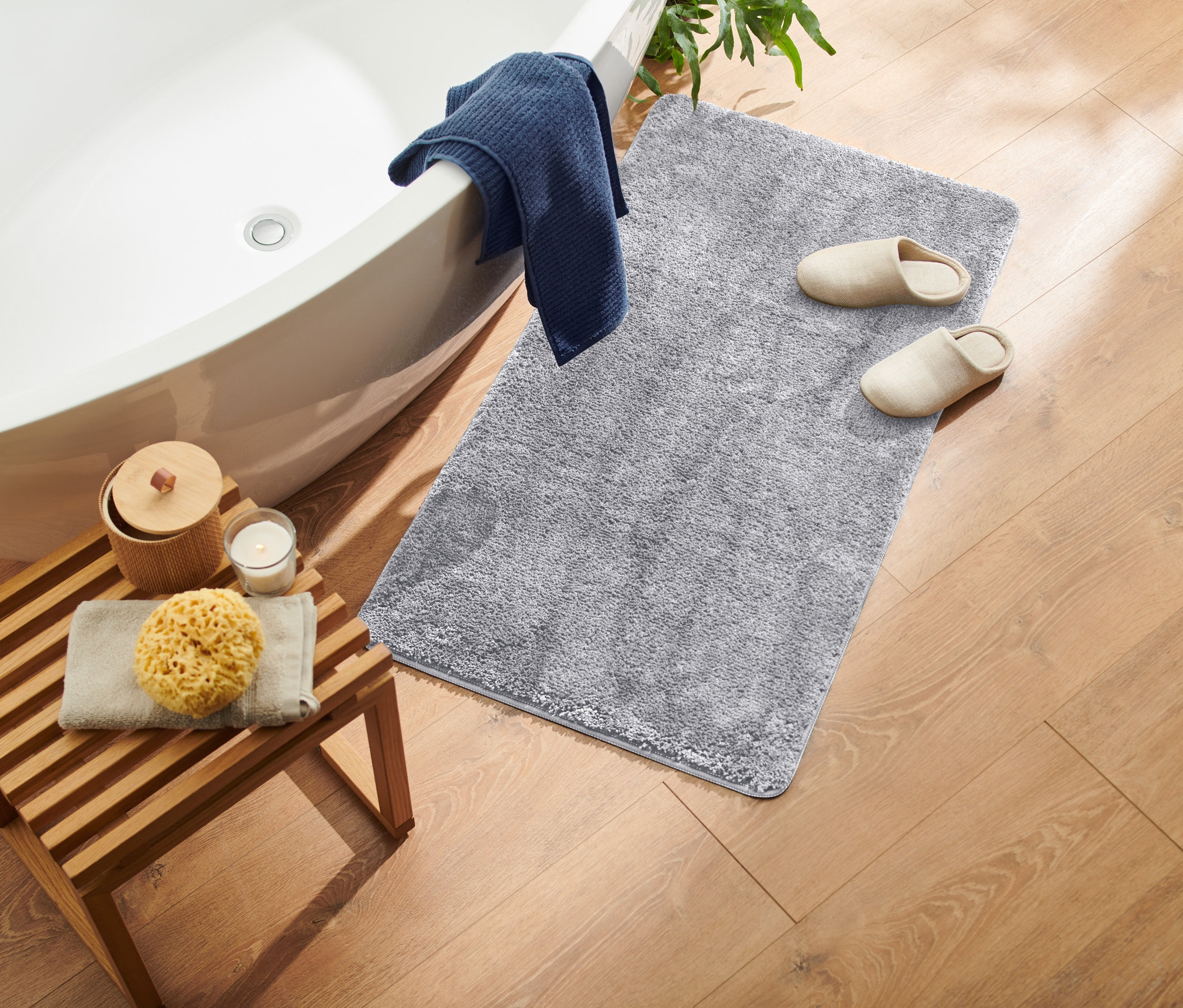 Küp, gri tüylü halı, bej terlikler, sünger ve mum bulunan ahşap tabure ile banyonun yukarıdan görünümü.