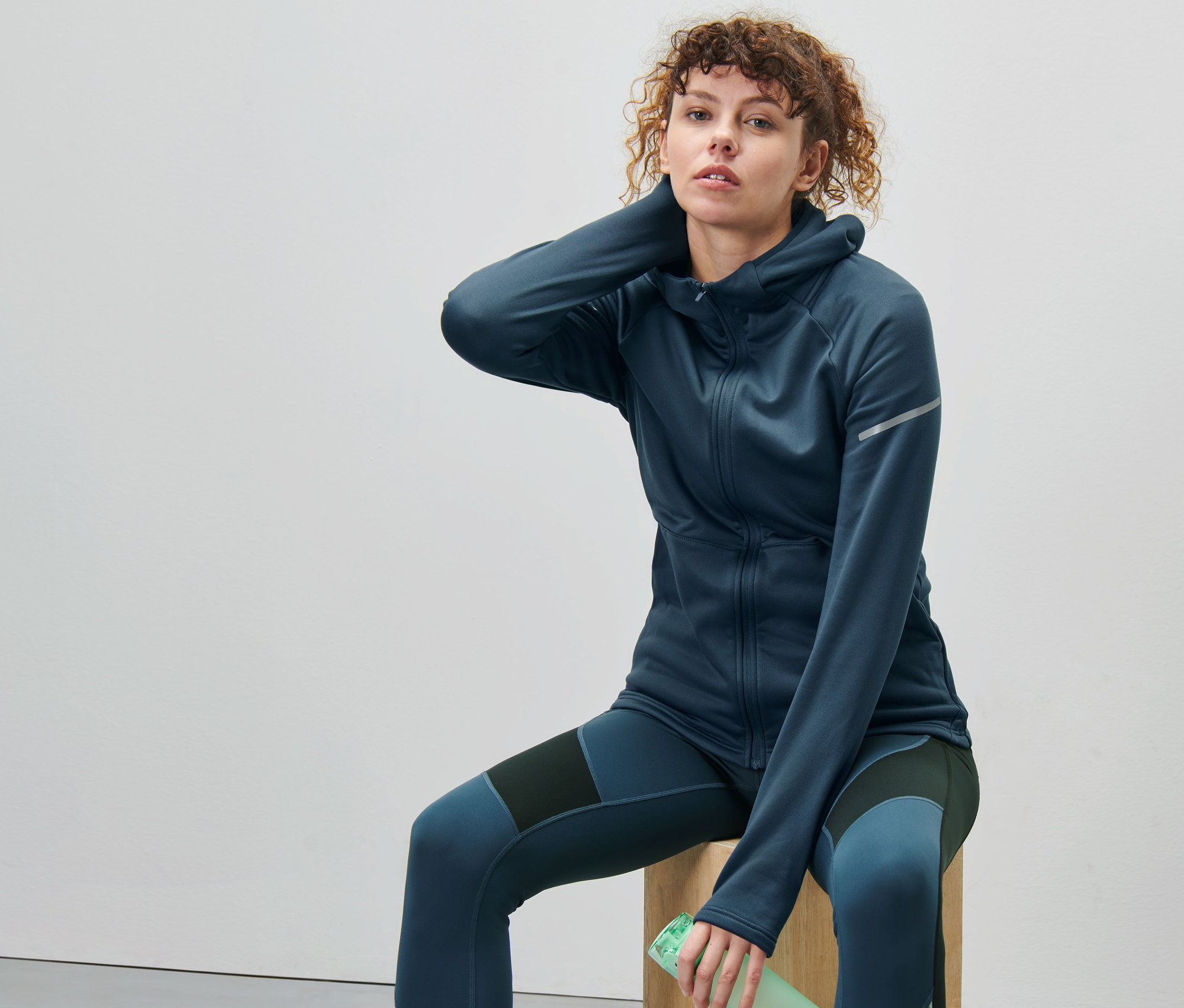 Kadın ahşap bir küpün üzerinde oturuyor, lacivert termal ceket giyiyor.