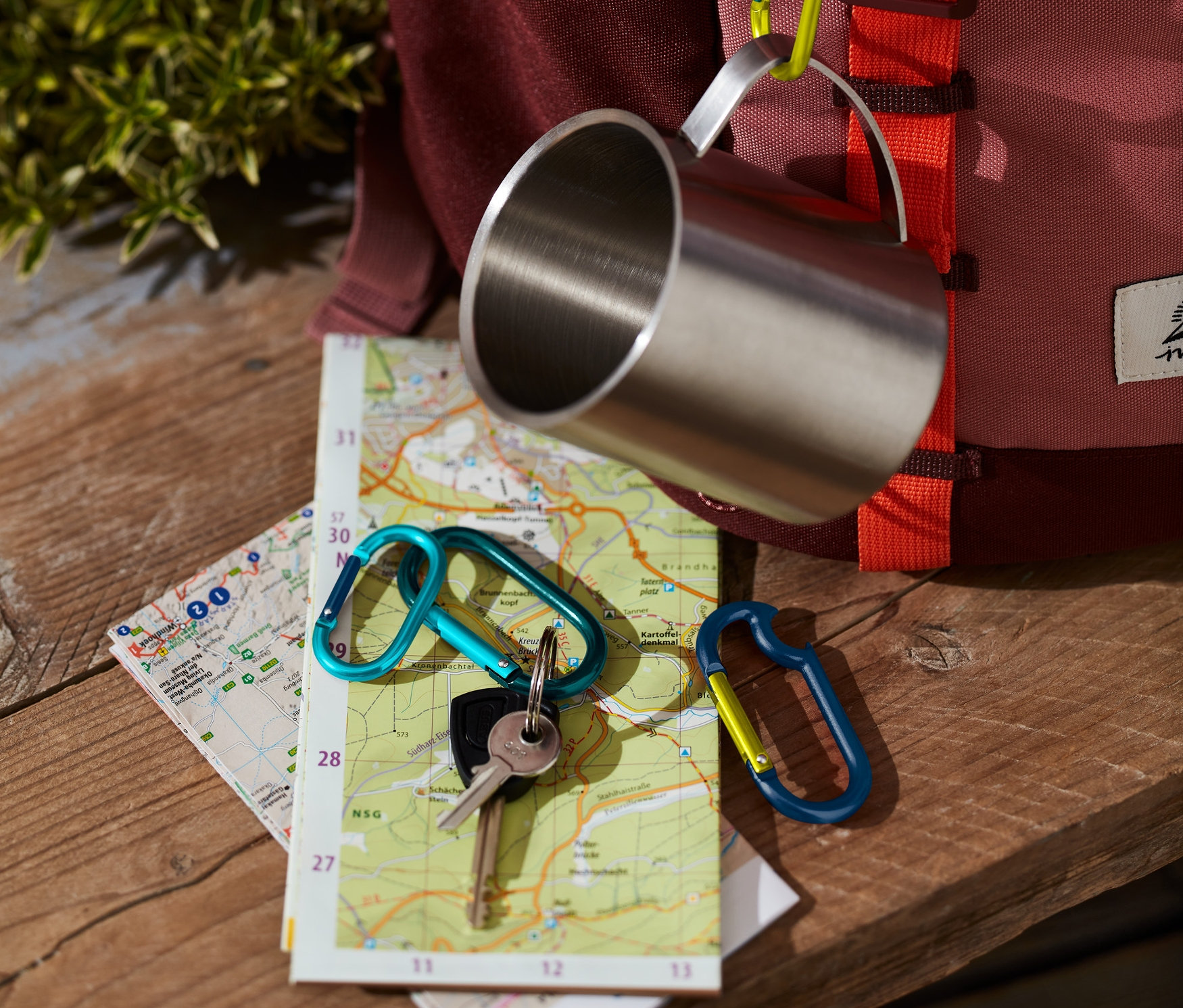 Paslanmaz çelik bir bardak, 4 adet karabina kancadan biriyle bir sırt çantasına asılıdır. Bir harita üzerinde başka bir karabina ile anahtarlar duruyor.