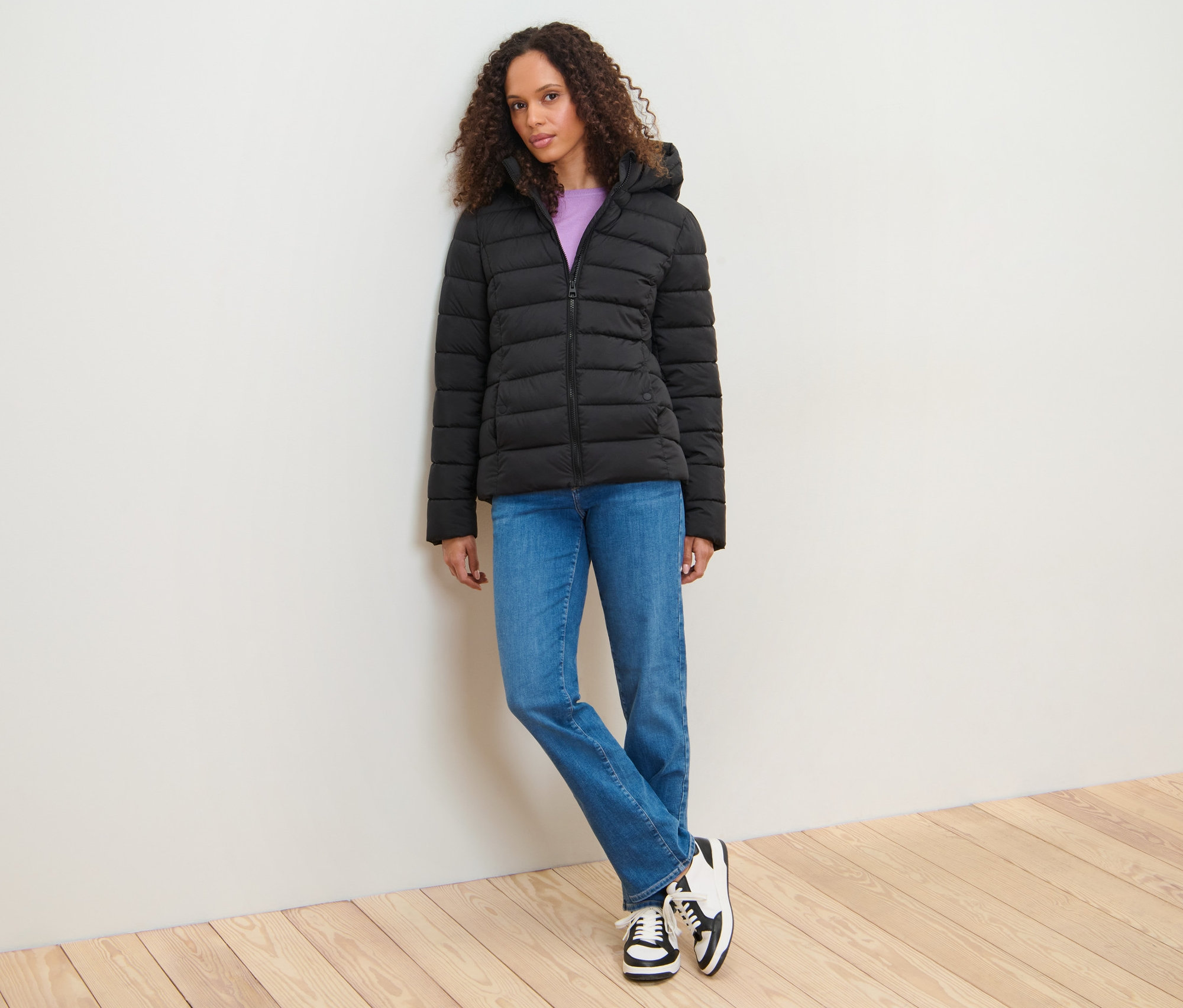 Duvara yaslanmış bir kadın kapüşonlu kapitone mont giyiyor.