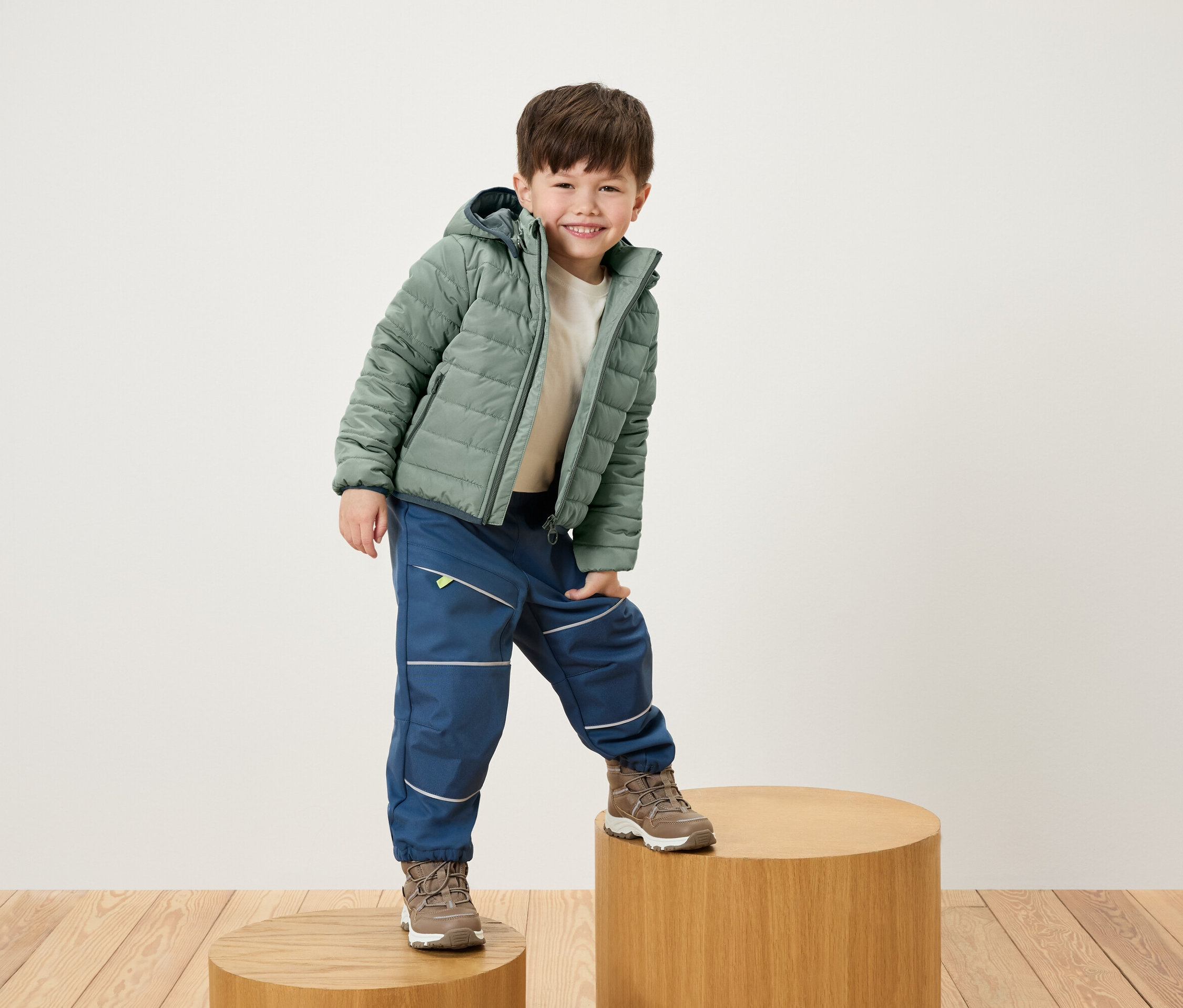 Gülümseyen çocuk, yeşil ceket, mavi pantolon ve kahverengi ayakkabılarla ahşap bir kaide üzerinde poz veriyor.