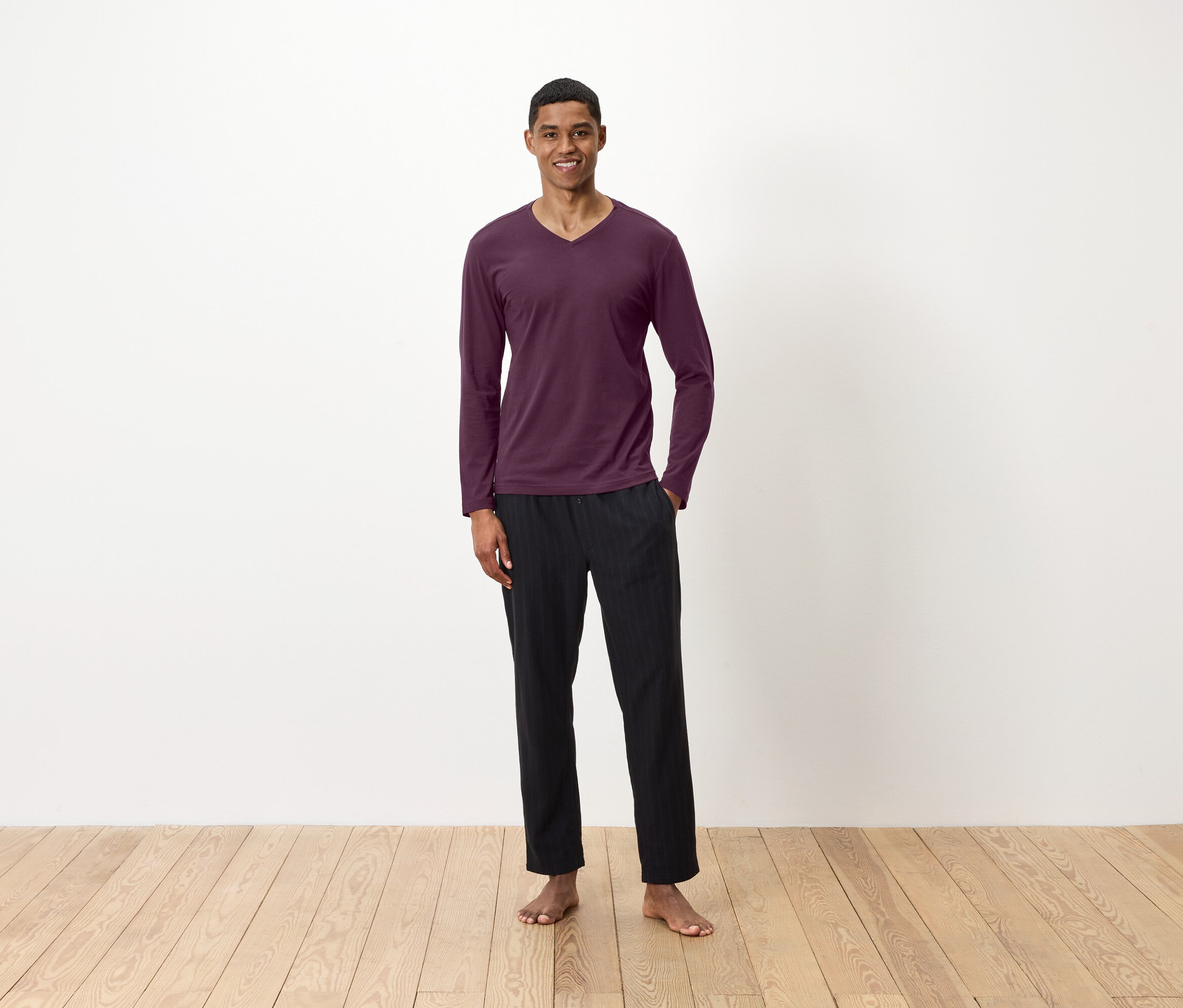 Bir adam ahşap zeminde çıplak ayakla duruyor, bordo uzun kollu bir tişört ve siyah pantolon giyiyor.