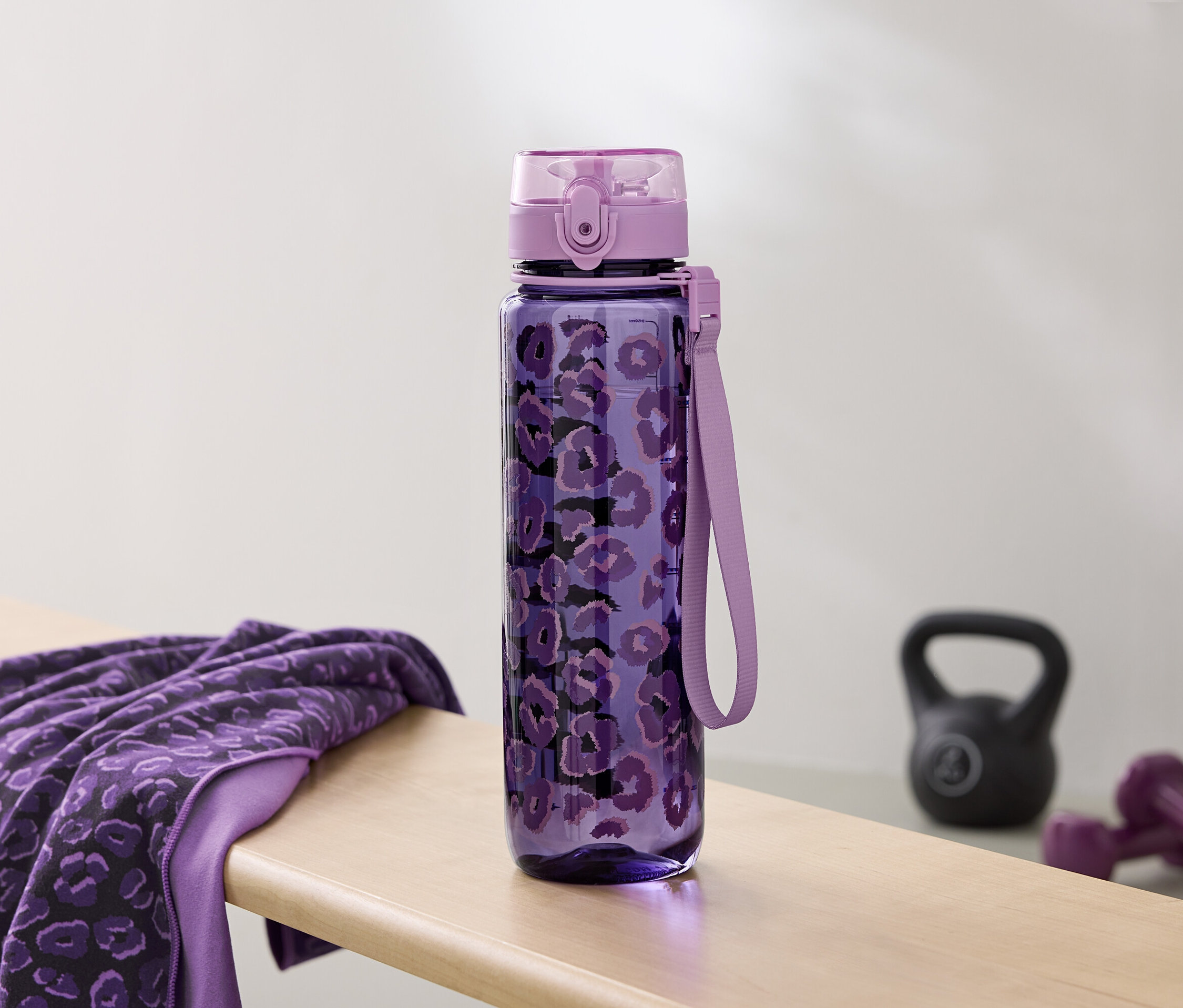 Desenli mor su şişesi, bir bankta havlu ve dambılların yanında duruyor.