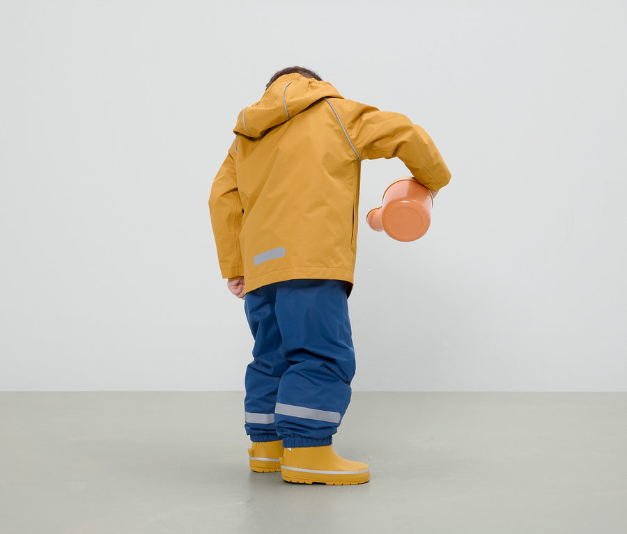 Hardal rengi bir ceket, mavi pantolon ve sarı çizme giyen bir çocuk, turuncu bir sulama kabı tutuyor.