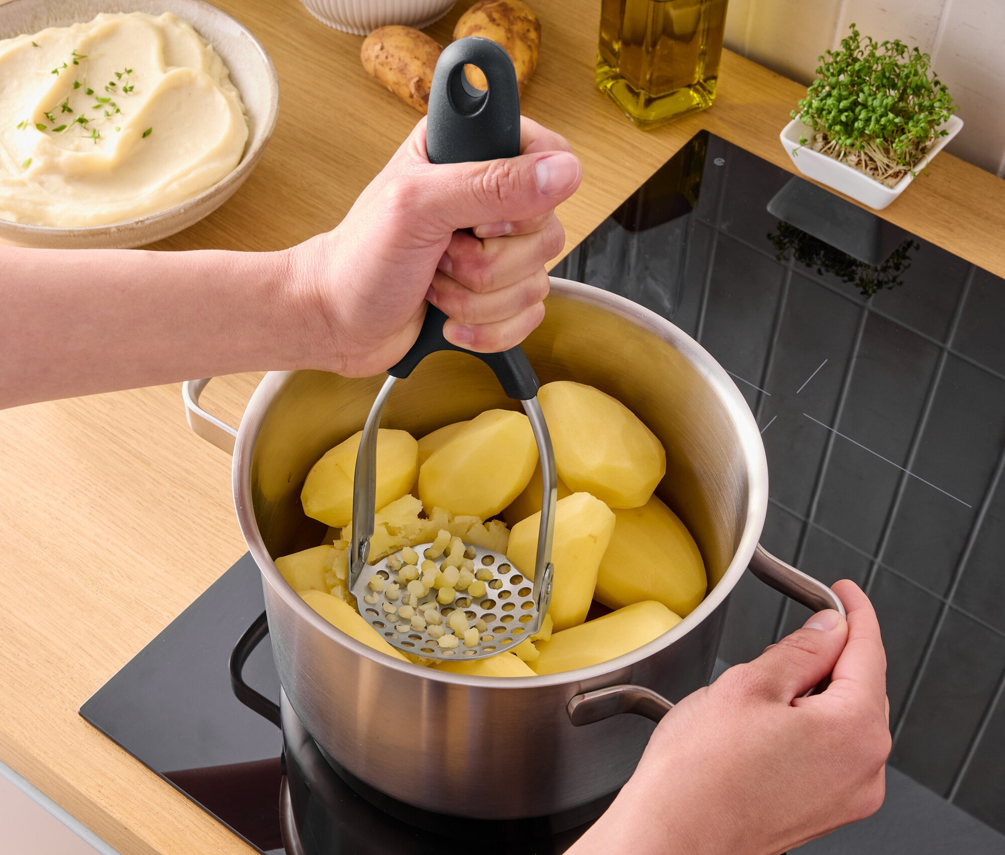 Bir kişi ocaktaki bir tencerede patates ezici kullanarak haşlanmış patatesleri eziyor.