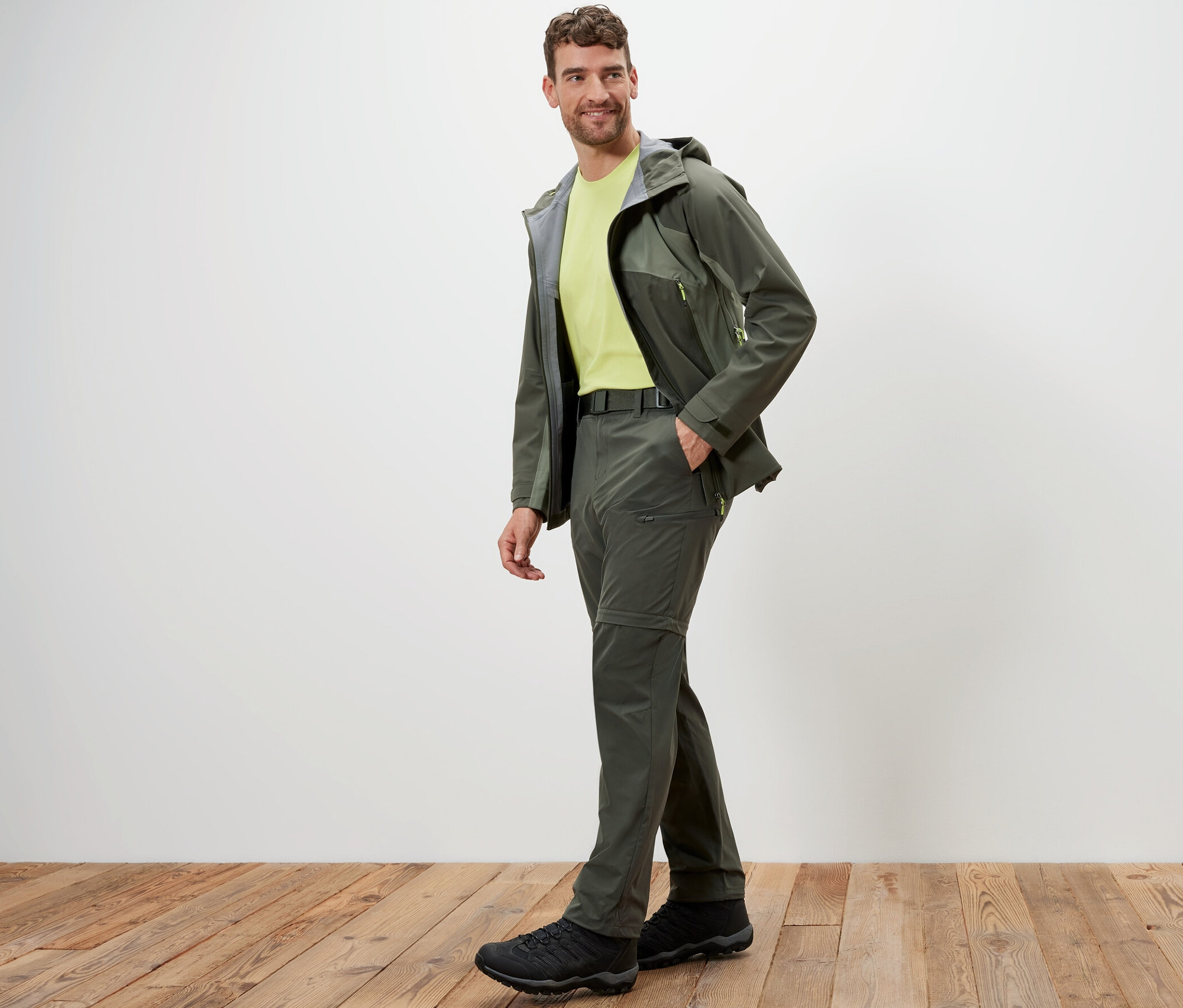 Adam ahşap zeminde haki 2'si 1 arada fonksiyonel pantolon ve ceketle poz veriyor.