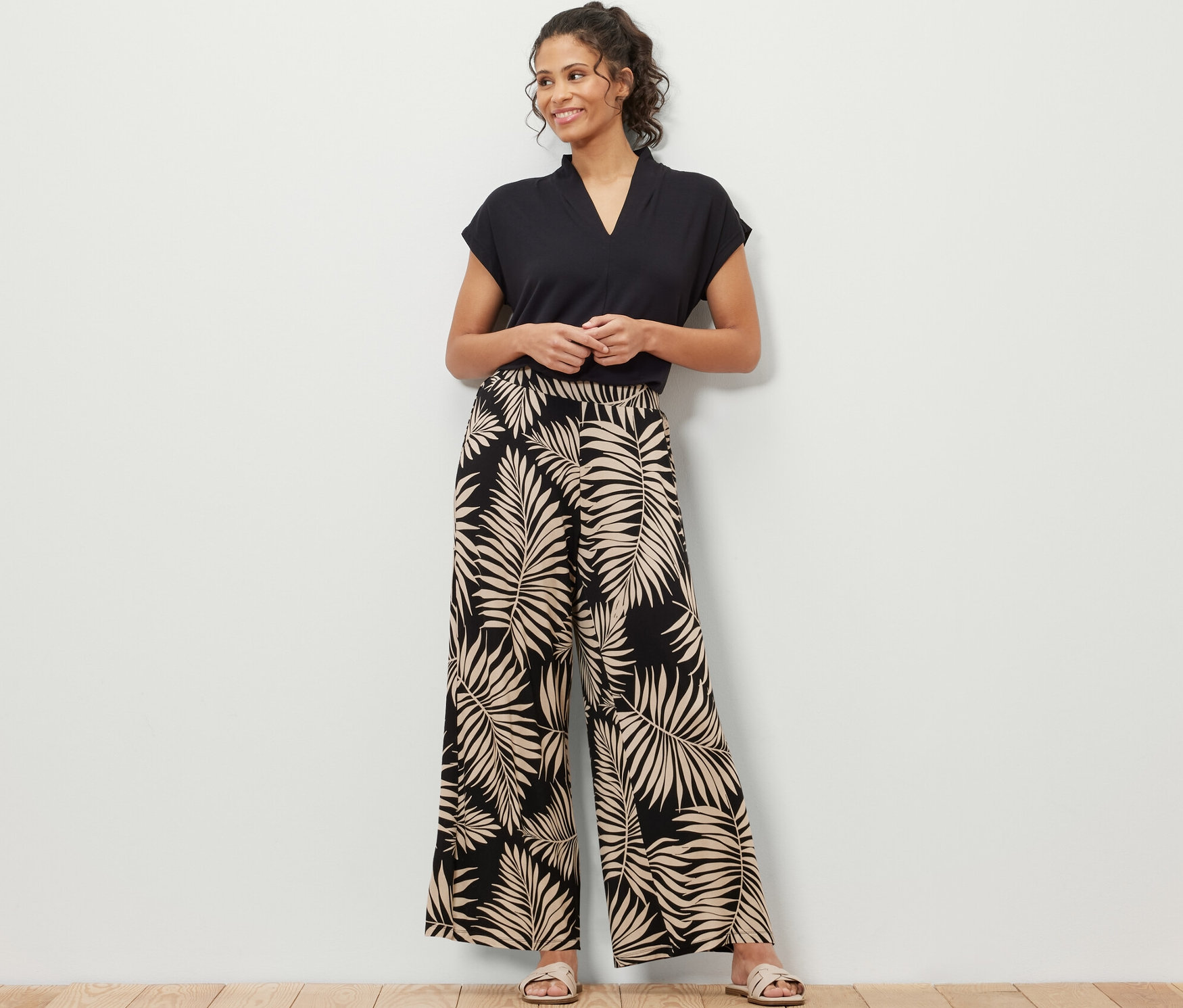Bir kadın ayakta duruyor, üzerinde siyah bir tişört ve yaprak desenli, baskılı bir palazzo pantolon var.
